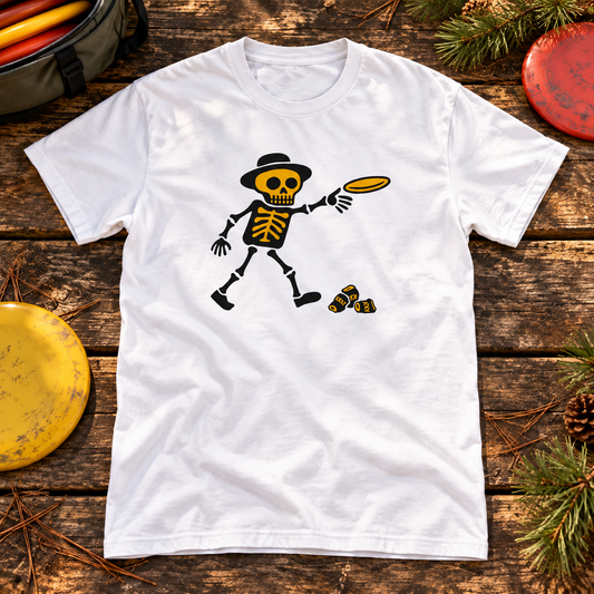 White Fribaaja skeleton disc golfer T-shirt on an outdoor wooden table with frisbee discs, a disc golf bag and pine sprigs.
