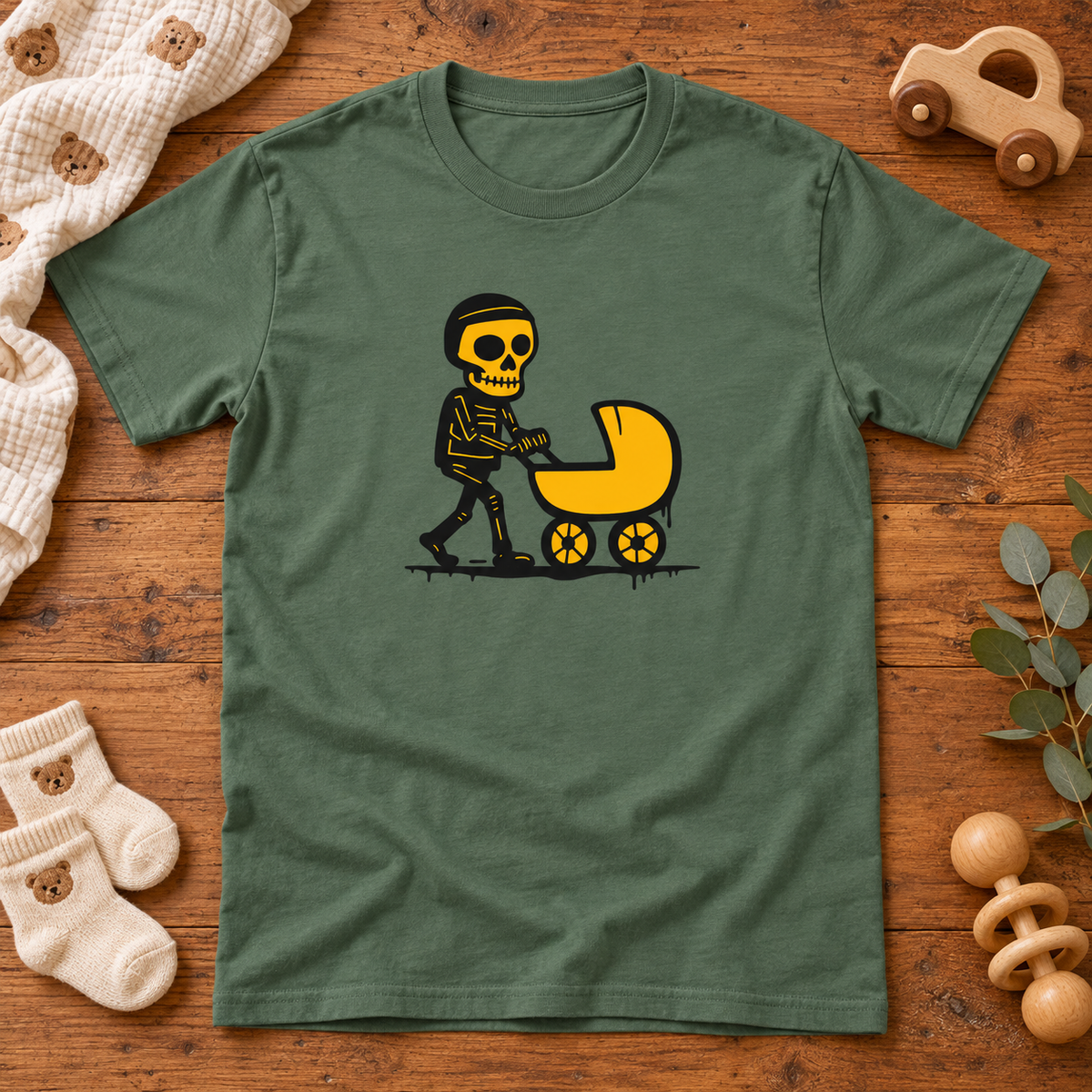 Military green Tuore Isä skeleton pushing a pram T-shirt on a nursery table with baby socks, blanket and wooden toy.